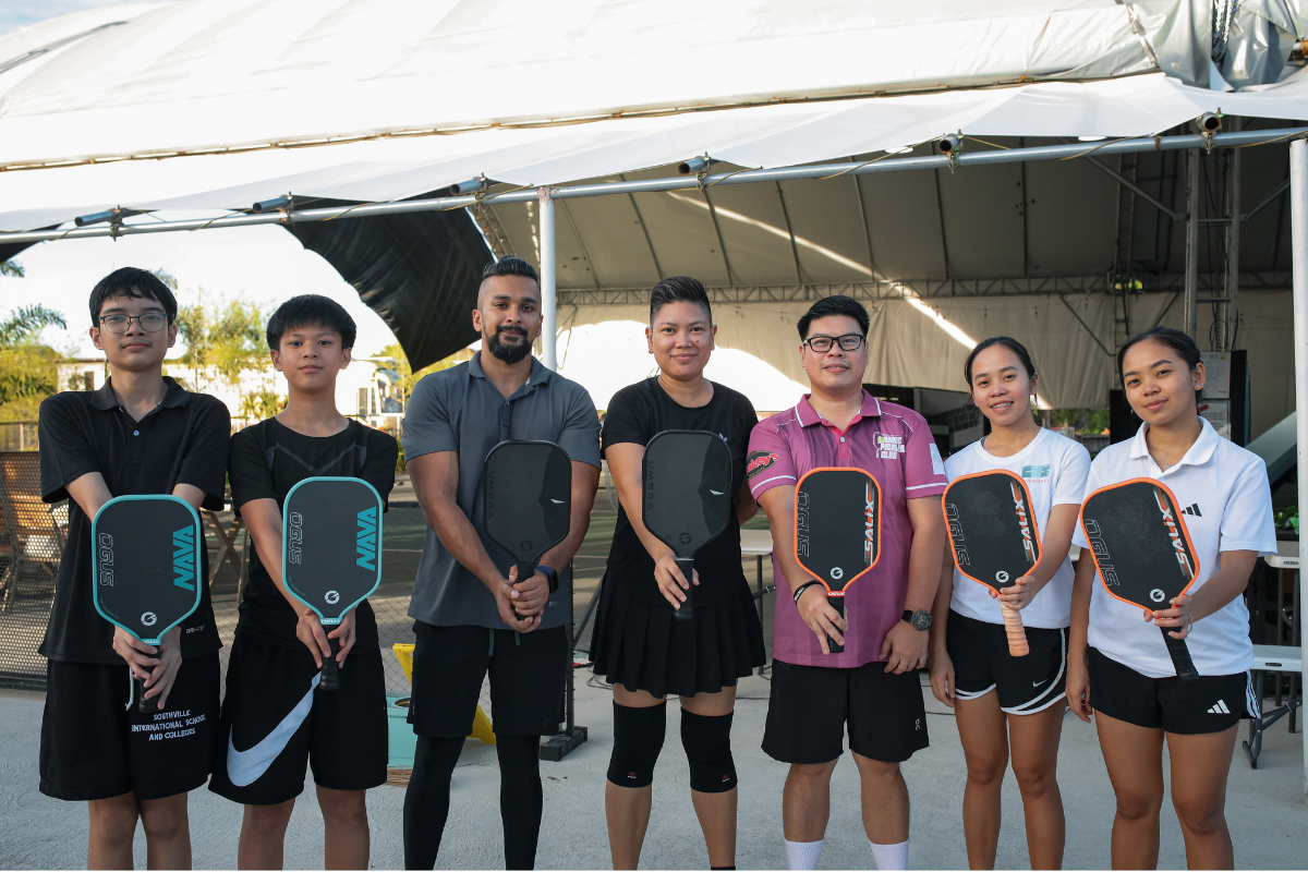 A group of seven people standing together under a white canopy tent, each holding a pickleball paddle. The group is dressed in athletic wear, and the paddles feature visible brands including PIP, Mamba, and Adidas.